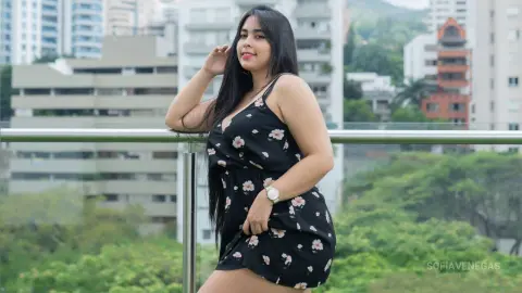Smiling woman in a black floral sundress posing on a city balcony overlooking lush green trees.