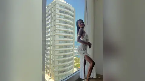Smiling Black woman in a white lace maxi dress posing by a high-rise window with a sunny city view.