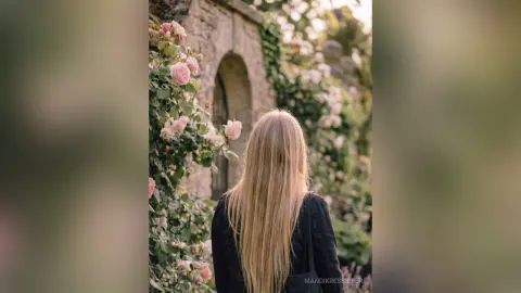Blonde woman in a black outfit standing in a romantic rose garden by a stone archway.