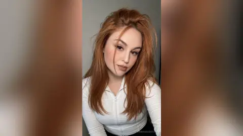 Redheaded woman in a white long-sleeve top posing with a soft, natural look in a minimalist indoor setting.