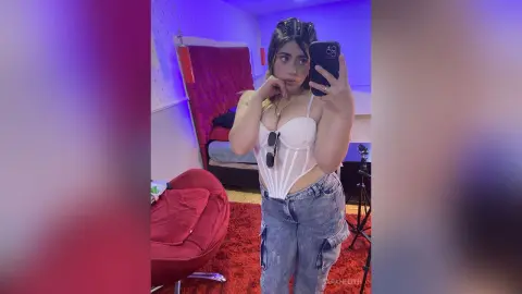 Young woman in a white corset top and baggy denim jeans taking a selfie in a bedroom with moody lighting.
