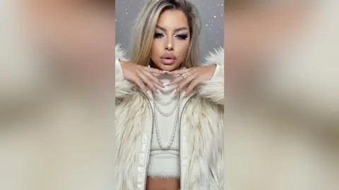 NicolePeach poses with her hands near her face, wearing a white furry jacket and a white top with a long pearl necklace.