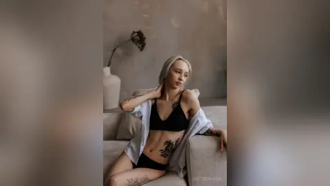 Tattooed woman in black lingerie and white shirt relaxing on a sofa in a minimalist, moody living room.
