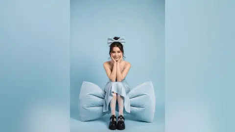 Cheerful young woman in a light blue dress sitting on a giant bow against a pastel blue background.