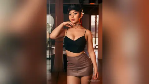 VickyPortman poses in a black bra top and brown leather skirt, looking at the camera with her hand near her neck.