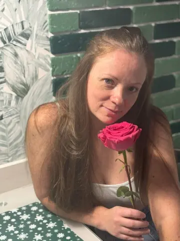 Smiling woman in a white tank top holding a single pink rose in a room with green brick walls.