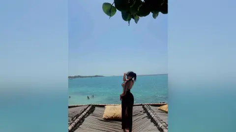 Woman in black bikini and trousers relaxing on a seaside net hammock overlooking a tropical turquoise ocean.
