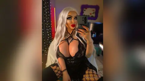 Glamorous woman with long white hair in a black lace outfit taking a selfie in a dimly lit room.