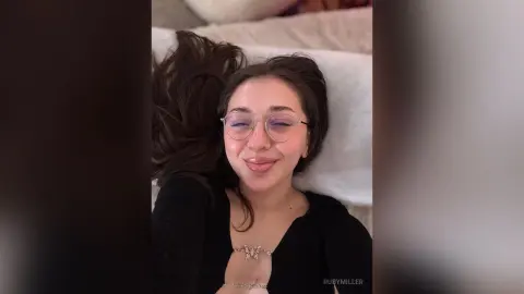 Smiling young woman with glasses in a black top relaxing on a white pillow in a cozy indoor setting.