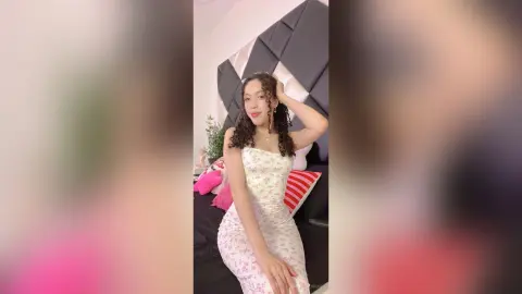 Young woman in a white floral sundress posing in a bright, modern bedroom with a cozy aesthetic.
