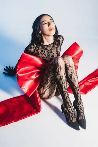 AilynHoney — AilynHoney sits on a white floor wearing a black lace bodysuit and a massive red satin bow.
