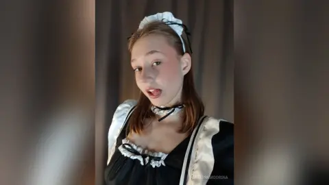 Young woman in a black and white maid costume with a frilled headband posing against a dark backdrop.