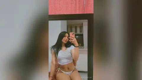 MarcelaMilan takes a mirror selfie wearing a tight white crop top and tiny white bikini bottoms, showing off her long dark hair and curves.