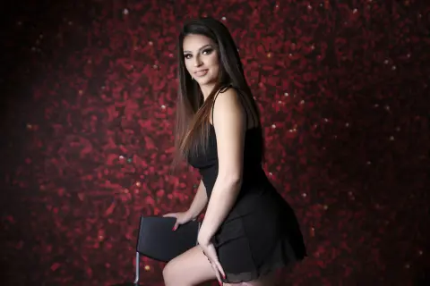 Elegant woman in a black cocktail dress posing against a glamorous red glitter background.