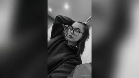 Moody black and white portrait of a young woman with glasses wearing a cozy dark sweatshirt indoors.