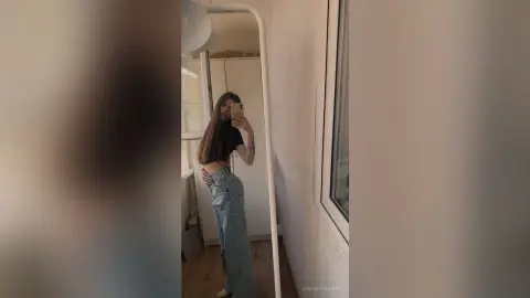 Young woman in a black crop top and baggy denim jeans taking a mirror selfie in a bright minimalist room.