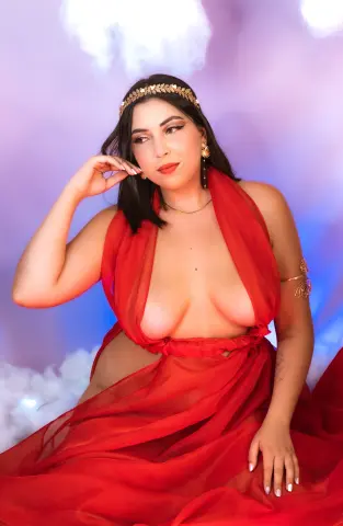 Woman in a flowing red goddess dress and gold laurel crown posing against a dreamy pink cloud background.