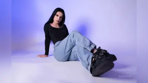 Young woman in black long-sleeve shirt and light blue jeans sitting against a purple studio background.