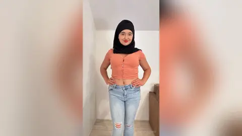 Smiling young woman in a black hijab and peach crop top posing in a bright modern room.