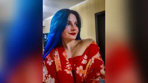 Woman with long blue hair wearing a red floral kimono in a warm indoor setting.