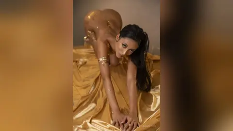 Glamorous woman with gold body glitter posing on shimmering satin fabric in a luxurious, glowing setting.