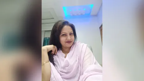 ManishaJi — ManishaJi smiles while wearing a light pink wrap and leaning against a white wall.