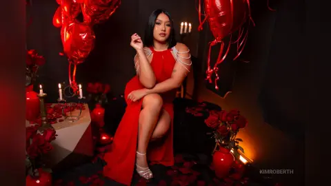KimRoberth poses in a red dress with silver chain strap detailing, sitting among red roses and red balloons.