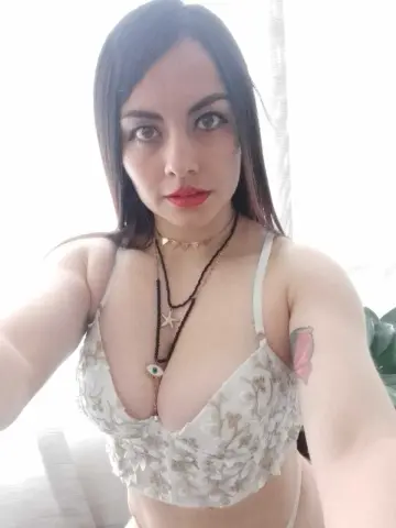 Young woman with long dark hair wearing a floral lace bralette and layered necklaces in a bright indoor setting.