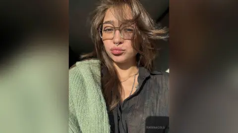 Young woman with glasses and nose ring wearing a dark shirt and green sweater in soft natural sunlight.