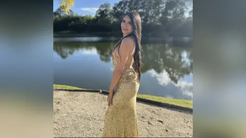 Smiling woman in a sparkling gold sequin evening gown posing by a serene lake at sunset.