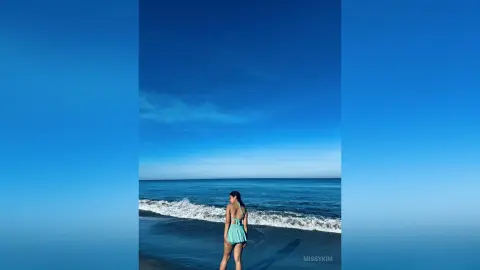 Young woman in a mint green swimsuit standing on a sunny beach facing the blue ocean waves.