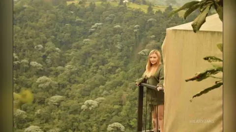 Blonde woman in an olive green dress standing on a balcony overlooking a lush, misty forest landscape.