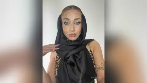 Woman in black silk headscarf and gold headpiece with glamorous makeup posing against a white background.