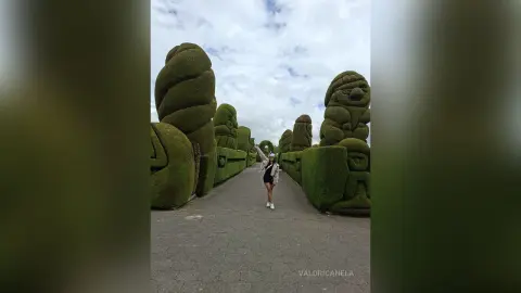 Stylish woman in a black outfit standing on a path between giant green topiary sculptures under a cloudy sky.