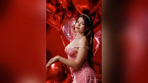 Young woman in a pink floral dress posing with red balloons in a romantic, festive red studio setting.
