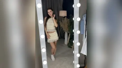 Young woman in a chic cream lace mini dress posing in a lighted vanity mirror for a stylish fashion look.
