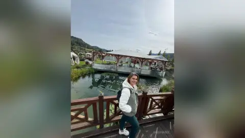 Smiling woman in a white hoodie and teal pants standing on a wooden deck by a scenic lake and pavilion.