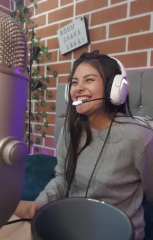 Smiling female streamer wearing a grey sweater and white headset in a cozy gaming room setup.