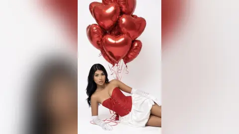 Glamorous woman in a red corset and white gloves posing with large red heart balloons against a white background.