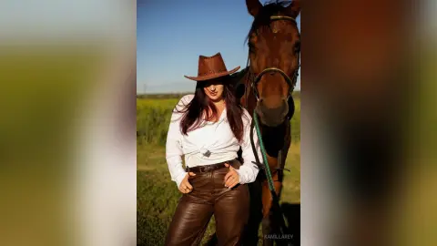 KamillaRey wears a brown cowboy hat and brown leather pants with a white long-sleeve shirt, posing next to a brown horse in a field.