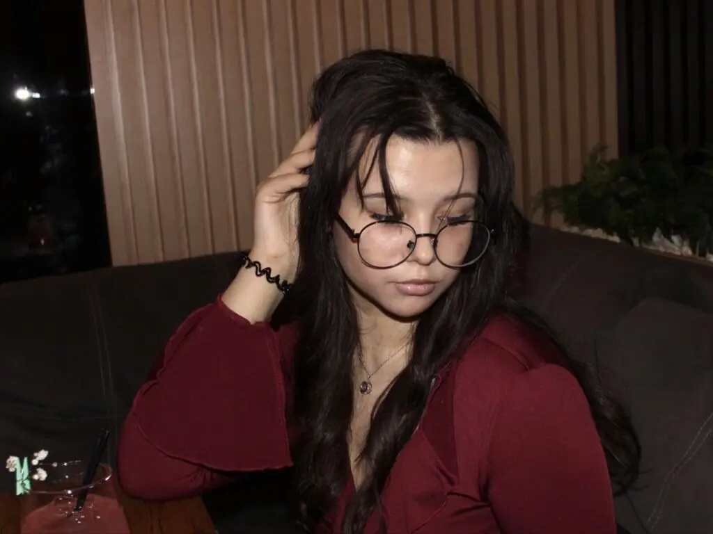Young woman with glasses in a red blouse sitting on a couch in a moody, dimly lit indoor setting.