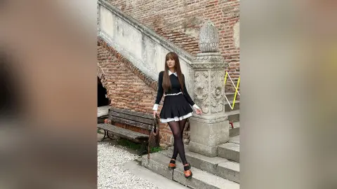 TiffanyAddams stands outdoors in a black and white schoolgirl-style dress with black tights and black shoes, posing next to a stone pillar and brick wall.