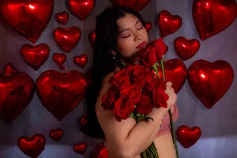 Romantic woman in pink holding a bouquet of red roses against a backdrop of red heart balloons.