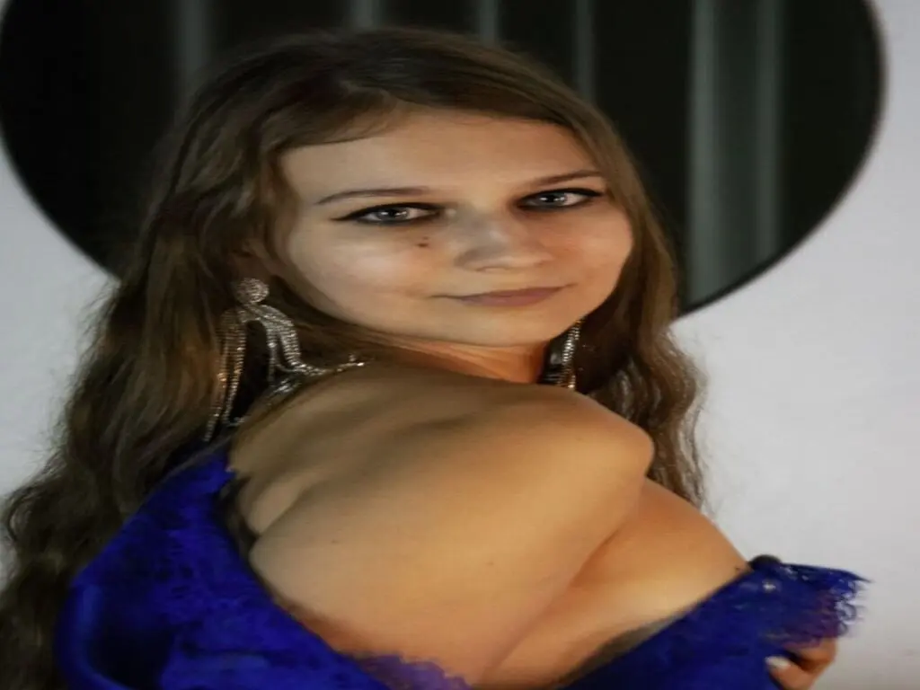Young woman with long wavy hair wearing a blue lace dress and silver earrings in a soft indoor setting.