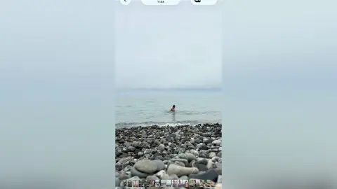 A person wading in the ocean waves on a misty, overcast rocky beach under a cloudy sky.