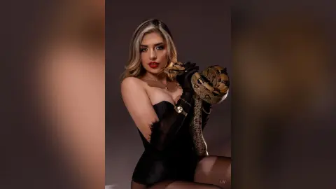 Glamorous woman in a black strapless outfit holding a large patterned snake against a dark studio background.