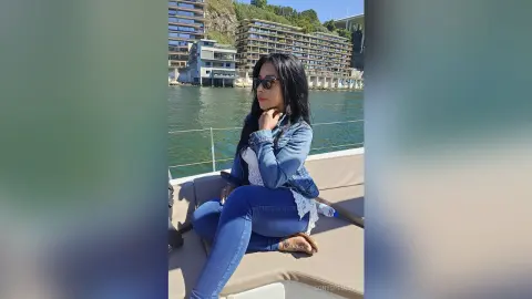Young woman in denim jacket and blue jeans relaxing on a boat in a sunny coastal harbor setting.