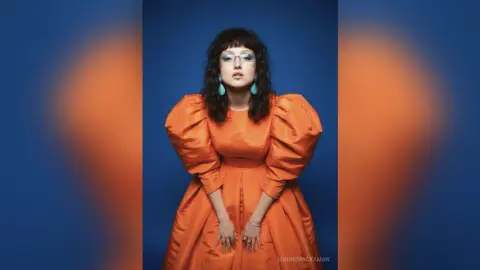 Stylish woman in an orange puff-sleeve dress and glasses posing against a blue background with a moody vibe.