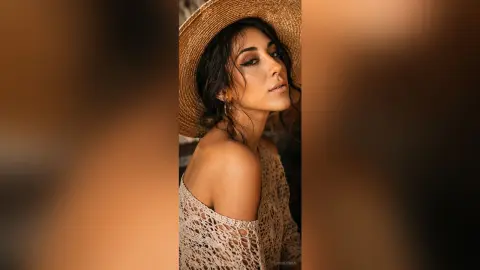 LorineSwan poses in a straw hat and a light, open-knit crochet top, looking over her shoulder with dark hair and heavy eye makeup.