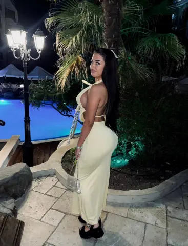 Woman in a cream backless maxi dress standing by a lit swimming pool at night.